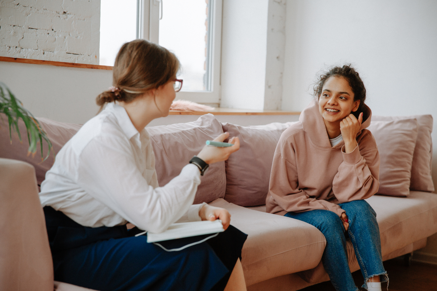 Therapist in a white shirt is sat on a sofa with a young person in a pink hoodie, talking.